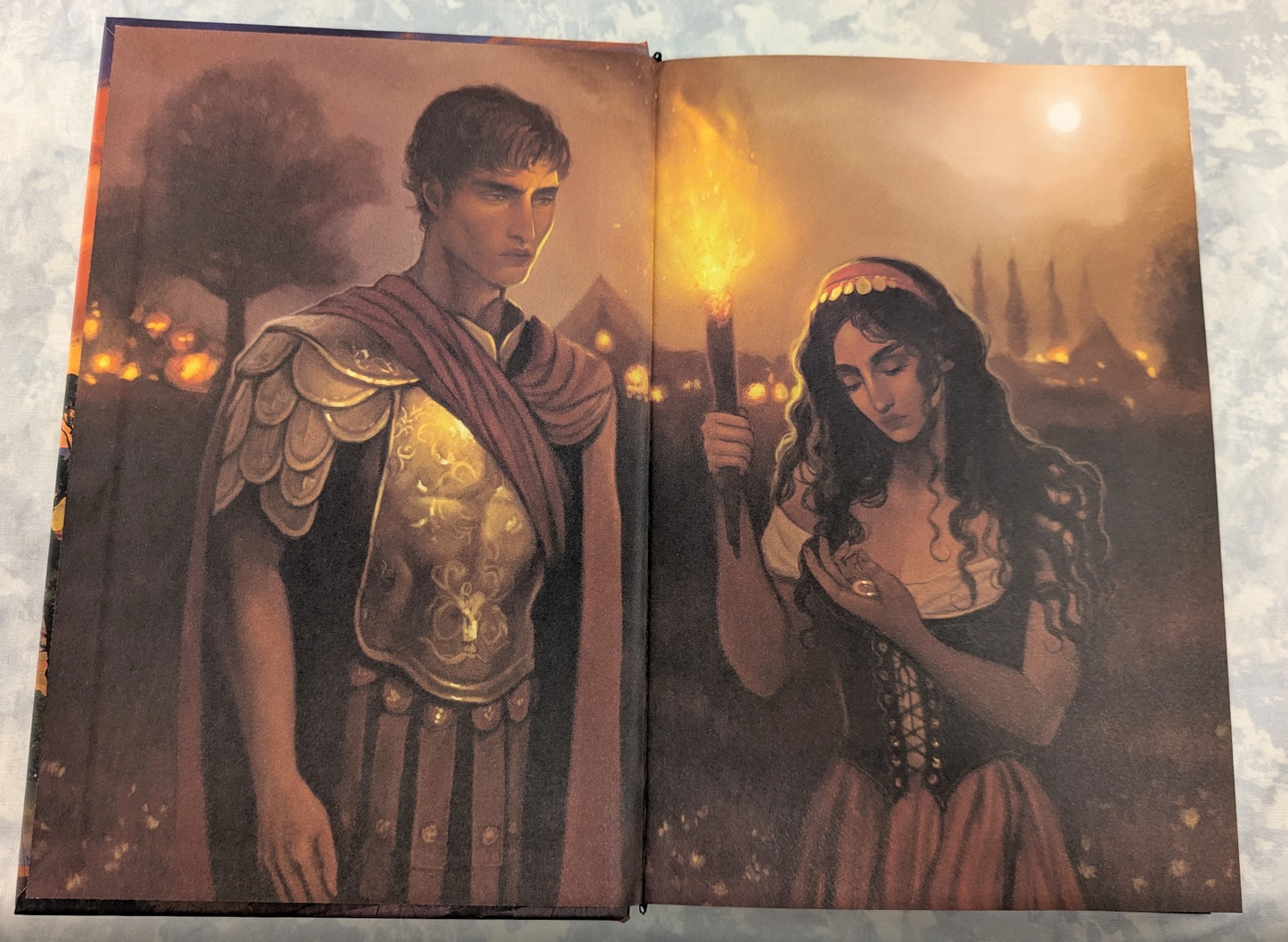 An open book showing two illustrated pages with a dark fantasy scene, featuring a woman holding a glowing torch and a figure in a golden robe, photographed against a light textured background.