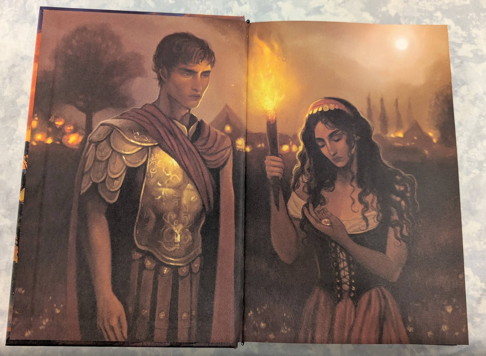 An open book showing two illustrated pages with a dark fantasy scene, featuring a woman holding a glowing torch and a figure in a golden robe, photographed against a light textured background.