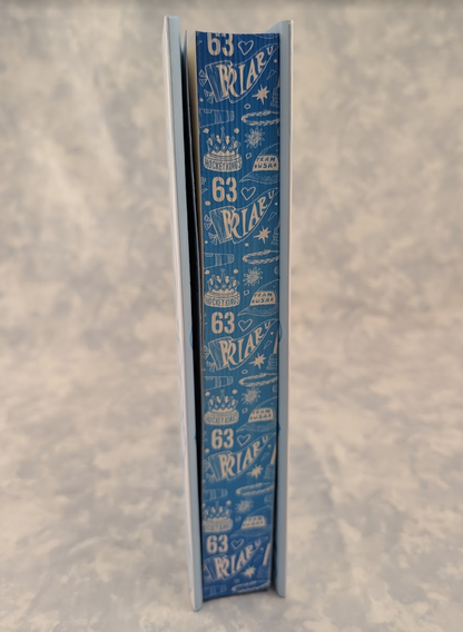 A tall, narrow book standing upright on a mottled light gray surface. The stenciled edges are covered in a repeating white pattern featuring the number 63 and small illustrated icons like cakes, stars, and bows.