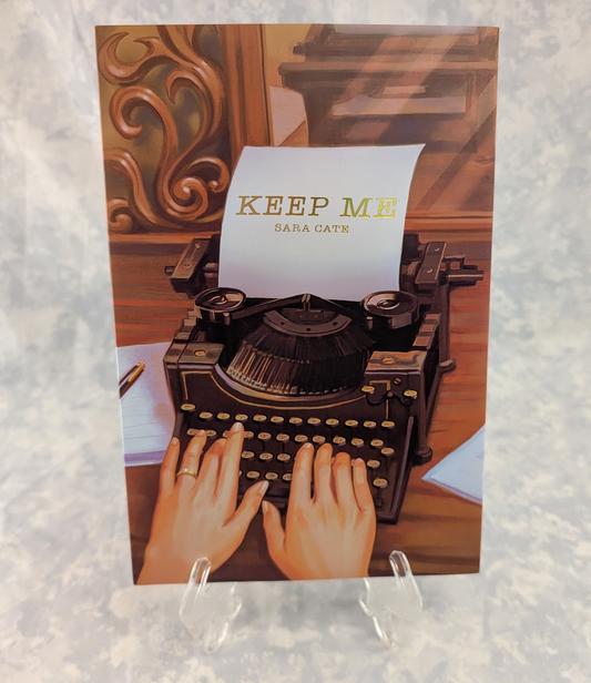 A hardcover book displayed on a small clear stand, showing an illustrated vintage typewriter with a paper sheet reading “KEEP ME” and “SARA CATE.” Hands are typing on the typewriter, set against a warm brown interior background.
