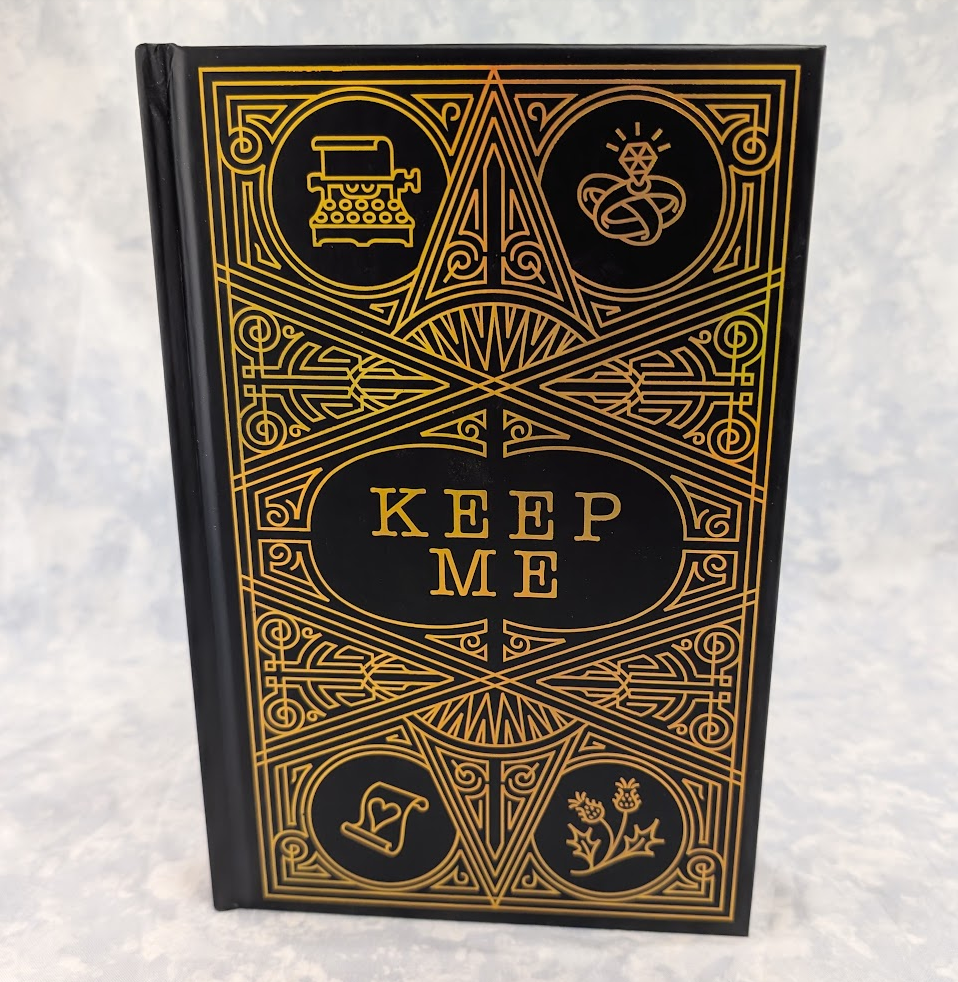 A hardcover book with a black cover and ornate gold Art Deco-style line art and typography. The center features the words 'KEEP ME' in gold, surrounded by decorative geometric illustrations.