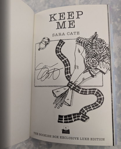 An open book showing a black-and-white autographed page with the title 'KEEP ME' and 'SARA CATE' on a white page, photographed against a light stone-like surface.