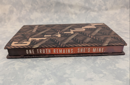 A hardcover book titled 'Knotted' with a dark brown textured cover featuring rope patterns and gold lettering, resting on a mottled gray surface. The spine reads 'ONE TRUTH REMAINS. SHE'S MINE' in capitol letters.