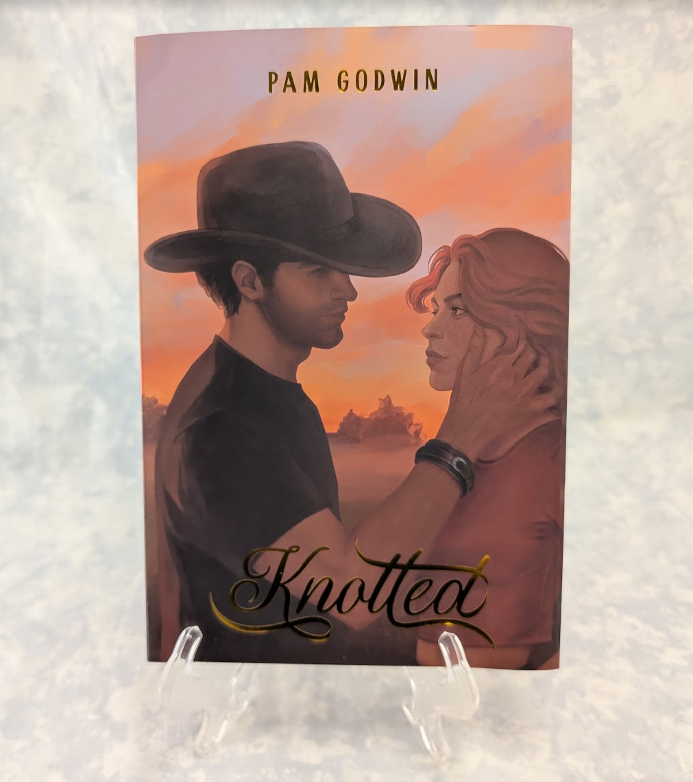 A book featuring an illustrated cowboy and woman at sunset, with the text 'PAM GODWIN' at the top and 'Knotted' at the bottom, displayed on clear stands against a light textured background.